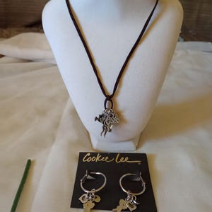 Pode incluir: Um colar de couro marrom com um pingente de prata com um desenho de uma menina com asas e corações. Brincos de argola combinando com o mesmo charme pendurados nos aros. Os brincos e o colar são embalados em um cartão preto com o texto "Cookee Lee" impresso em branco.