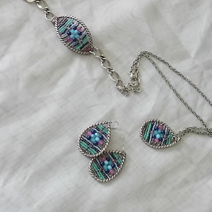 Puede incluir: Un conjunto de joyas que incluye una pulsera, un collar y pendientes. Cada pieza tiene un dise&ntilde;o ovalado con un marco de metal plateado y est&aacute; llena de cuentas de colores en tonos azules, rosas y verdes.