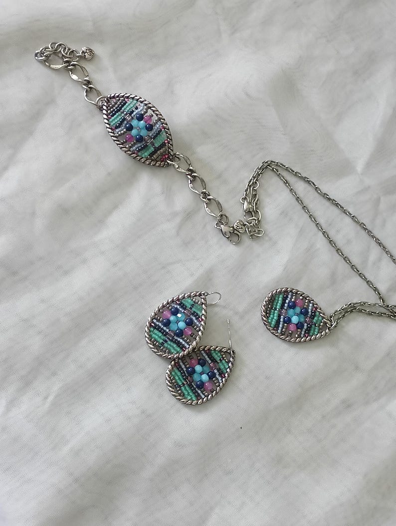 Puede incluir: Un conjunto de joyas que incluye una pulsera, un collar y pendientes. Cada pieza tiene un dise&ntilde;o ovalado con un marco de metal plateado y est&aacute; llena de cuentas de colores en tonos azules, rosas y verdes.
