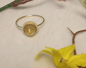 Puede incluir: Anillo dorado con una gema redonda de color marrón claro. La gema está rodeada por un borde decorativo con cuentas. El anillo se encuentra sobre un fondo blanco suave con detalles florales.