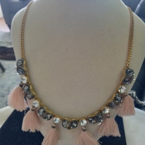 Op de afbeelding: Een gouden ketting met een rij gefacetteerde, donkergrijze en heldere kristallen. De ketting heeft een franje van roze kwastjes.