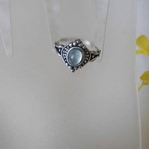 May include: A silver ring with an oval, light blue gemstone at the center. The ring has a decorative, beaded design around the gemstone and on the band. The ring is displayed on a clear, plastic hand.