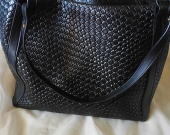 May include: Black woven leather tote bag with double handles and a shoulder strap. The bag has a rectangular shape and a textured surface. The bag is on a white surface.