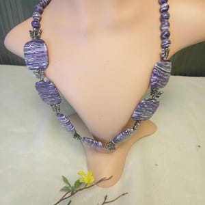 Puede incluir: Collar de cuentas veteadas en morado y blanco, expuesto en un maniquí. El collar presenta cuentas rectangulares y redondas, conectadas por eslabones plateados con forma de mariposa. Una pieza de joyería llamativa, ideal para añadir color.