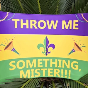 Op de afbeelding: Een groene, gele en paarse Mardi Gras-gooiflag met de tekst "THROW ME SOMETHING, MISTER!!" en een fleur-de-lis-ontwerp.