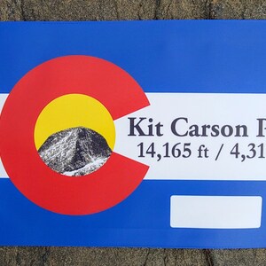 May include: A blue and white flag with the Colorado state flag in the center. The flag has the text "Kit Carson Peak 14,165 ft / 4,317 m" printed on it.