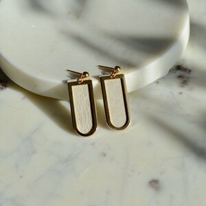 May include: A pair of gold-toned earrings with a white leather rectangle insert. The earrings have a simple, modern design.