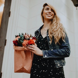 May include: A woman wearing a black and white patterned dress and a blue denim jacket holds a bouquet of red, pink, and white flowers wrapped in brown paper.