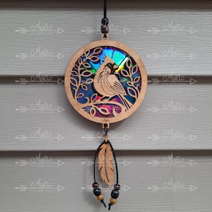 Könnte beinhalten: Vogel-Suncatcher aus Holz mit einem lasergeschnittenen Kardinal-Design. Der Kardinal sitzt auf einem Ast in einem kreisförmigen Rahmen mit Blattausschnitten. Ein farbenfrohes, schillerndes Material füllt die Mitte. Eine Holzfeder und Perlen hängen darunter.