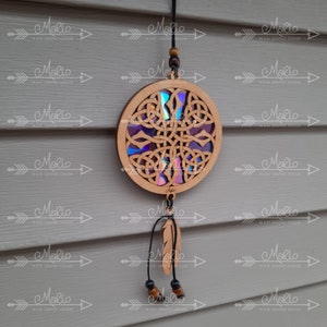 May include: A wooden dreamcatcher with a circular, intricate design featuring a Celtic knot pattern. The knotwork is filled with iridescent holographic film. A single black feather hangs from the bottom of the dreamcatcher.