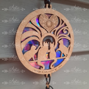 May include: A circular wooden ornament with a rainbow iridescent background. The ornament features a heart design with a mandala pattern and three cats silhouetted against the background. The text "MADE IN KELOWNA BC CAN" is printed on the bottom of the ornament.