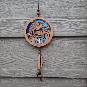 May include: A wooden dreamcatcher with a circular design featuring two dolphins and waves. The dreamcatcher is decorated with iridescent blue and green paint and has a black feather hanging from the bottom.