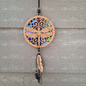 May include: A wooden dreamcatcher with a blue and green iridescent dragonfly design. The dreamcatcher has a black cord with brown beads and a wooden feather hanging from it.