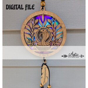 Può includere: Decorazione murale circolare in legno con un design a cobra tagliato al laser, foglie e sfondo iridescente. Le parole "DIGITAL FILE" sono in alto. Una piuma di legno e perline sono appese sotto.