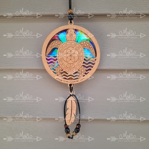 Op de afbeelding: Een houten schildpad-zonenvanger met een regenboogkleurige, iriserende afwerking. De schildpad is omringd door golven en heeft een levensbloem-ontwerp in het midden. De zonenvanger hangt aan een zwart koord met een bruine veer en kralen.