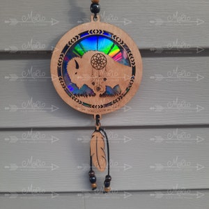 May include: A wooden dreamcatcher with a circular design featuring a buffalo silhouette and a rainbow iridescent background. The dreamcatcher has a brown leather cord and a single brown feather hanging from it.