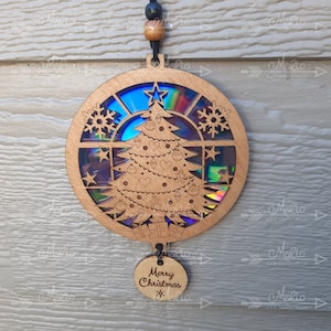May include: Wooden Christmas ornament with a laser-cut design featuring a Christmas tree, snowflakes, and stars. The ornament has a holographic backing that reflects multiple colors. A small, round tag at the bottom reads "Merry Christmas".