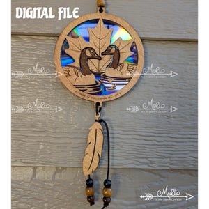 May include: Wooden ornament featuring a circular design with two geese swimming in water, beneath a maple leaf. The ornament includes a feather and beads, with the text "DIGITAL FILE" at the top. The text "MADE IN KELOWNA BC, CA" is also visible.
