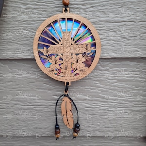 May include: A wooden circular ornament with a cross and lilies inside a rainbow iridescent background. The ornament is hanging from a black cord with a brown feather and beads.