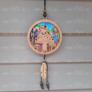 May include: A wooden circular ornament with a frog sitting on a mushroom. The ornament has a holographic iridescent background and is hanging from a black cord with brown beads and a wooden feather charm.