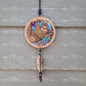 May include: A wooden sloth hanging ornament with a rainbow iridescent background and a brown feather hanging from the bottom. The ornament is round and has a black cord.
