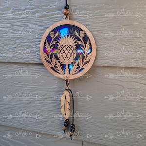 May include: A circular wooden ornament featuring a laser-cut design of a thistle flower. The ornament has a holographic iridescent background and hangs from a black cord with a wooden bead and a wooden feather charm.