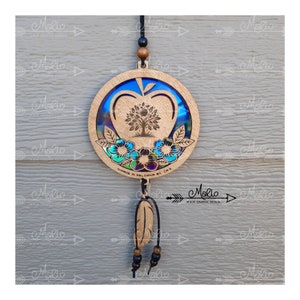 May include: A wooden apple-shaped ornament featuring a tree of life design within. The apple is decorated with iridescent blue and green colours and has a small feather hanging from it. The ornament is made in Kelowna, BC, Canada.
