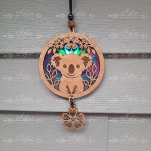 May include: A wooden ornament featuring a koala bear with a rainbow iridescent background. The ornament is hanging from a string and has a small wooden flower charm attached.