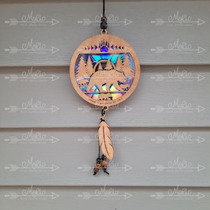 Puede incluir: Atrapasueños de madera con un diseño de oso y árboles. El atrapasueños está decorado con papel de aluminio iridiscente y tiene una pluma marrón colgando de él. El atrapasueños está colgado en una pared de madera marrón claro.
