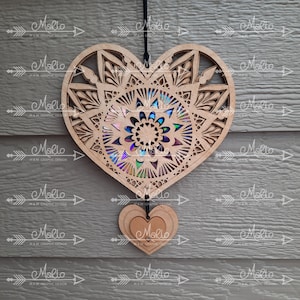 May include: A wooden heart-shaped ornament with a mandala design and iridescent accents. The heart is hanging from a black string and has a smaller heart-shaped ornament hanging from it. The smaller heart has the word "Marlo" written on it.