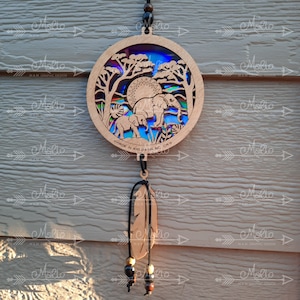 May include: A wooden circular ornament with a cutout design of elephants in a jungle scene. The ornament is hanging from a black cord with a brown feather and beads.