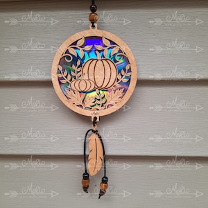 May include: A round wooden ornament with a holographic background featuring a carved design of pumpkins and leaves. A brown feather hangs from the bottom of the ornament with brown beads.