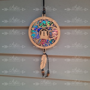 May include: A wooden circular dreamcatcher with a holographic iridescent design featuring two cats sitting on a branch with a heart between them. The dreamcatcher has a brown leather cord with a brown feather and brown beads hanging from it.