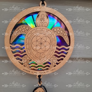 K&ouml;nnte beinhalten: Ein Holzausschnitt einer Meeresschildkr&ouml;te mit einer holografischen Scheibe in der Mitte. Die Schildkr&ouml;te ist von Wellen umgeben und hat ein Lebensblume-Design in der Mitte. Die Scheibe ist irisierend und reflektiert Licht in einem Regenbogen von Farben. Die Worte "MADE IN KELOWNA BC CA" sind auf der Unterseite des Ausschnitts geschrieben.