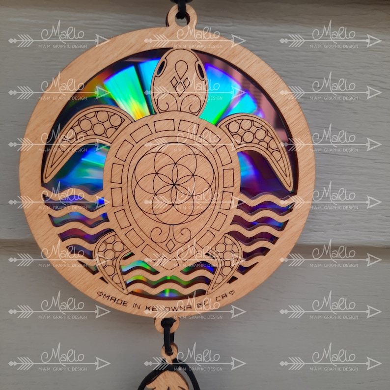 K&ouml;nnte beinhalten: Ein Holzausschnitt einer Meeresschildkr&ouml;te mit einer holografischen Scheibe in der Mitte. Die Schildkr&ouml;te ist von Wellen umgeben und hat ein Lebensblume-Design in der Mitte. Die Scheibe ist irisierend und reflektiert Licht in einem Regenbogen von Farben. Die Worte "MADE IN KELOWNA BC CA" sind auf der Unterseite des Ausschnitts geschrieben.