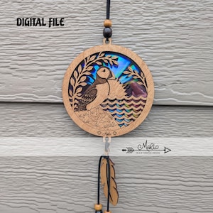 May include: A circular wooden hanging ornament featuring a laser-cut puffin standing on a rocky shore with a tree and sun, set against an iridescent background. The text DIGITAL FILE is visible at the top, and a wooden feather hangs below.