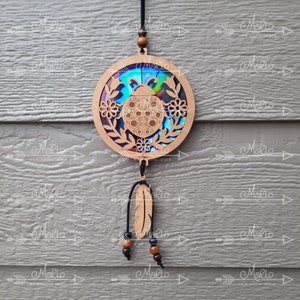 May include: A wooden ladybug dreamcatcher with a iridescent background and a feather hanging from the bottom. The dreamcatcher is decorated with small flowers and leaves.