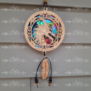 May include: A wooden dreamcatcher with a circular design featuring a dragon with iridescent wings. The dreamcatcher has a brown leather cord with a feather and beads hanging from it. The text "MADE IN KELOWNA BC CA" is printed on the wooden circle.