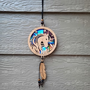 May include: A wooden unicorn dreamcatcher with a blue and purple iridescent background. The dreamcatcher is round and has a feather hanging from it. The text "MADE IN KELOWNA BC, CAN" is printed on the dreamcatcher.