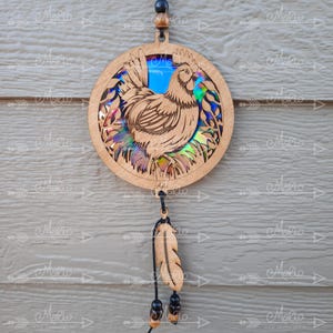 May include: Wooden circular ornament with a laser-cut design of a chicken in a leafy setting. The ornament has a iridescent blue and green background and a black cord with a wooden feather and beads hanging from it.