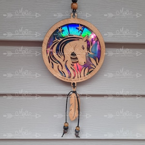 May include: Wooden dreamcatcher with a rainbow iridescent unicorn head cutout and stars. The dreamcatcher is hanging from a brown cord with a brown feather and beads.