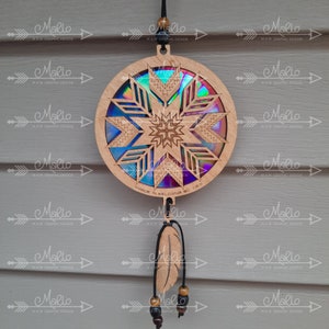 May include: A wooden dreamcatcher with a circular design featuring a starburst pattern and iridescent accents. The dreamcatcher has a feather and beads hanging from it. The text "TMROK KELOWNR RCCB" is visible on the back of the dreamcatcher.