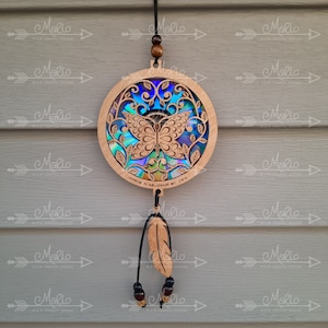 May include: A wooden circular dreamcatcher with a butterfly design and iridescent blue, green, and purple accents. The dreamcatcher is hanging from a black cord with brown beads and a wooden feather charm. The text "MADE IN KELOWNA BC CANADA" is printed on the dreamcatcher.