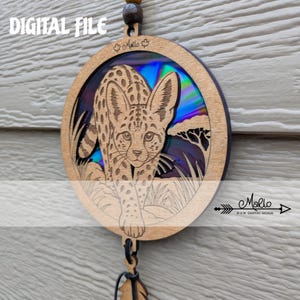 Puede incluir: Un adorno circular de madera con un diseño de serval cortado con láser sobre un fondo de arcoíris iridiscente vibrante. La pieza incluye una cuenta de madera oscura en la parte superior y un pequeño dije de madera en forma de pluma abajo.