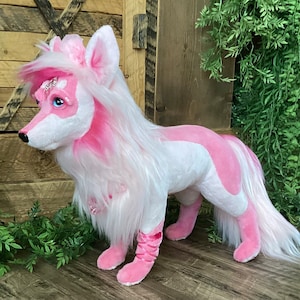May include: A pink and white plush toy dog with long white fur and a pink tail. The dog has a pink nose, blue eyes, and a sparkly pink crown on its head.