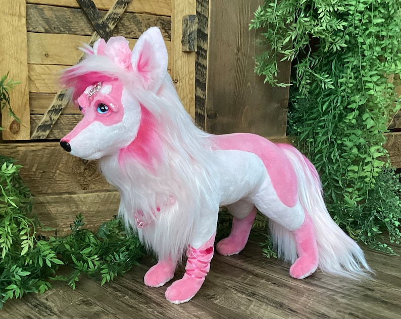May include: A pink and white plush toy dog with long white fur and a pink tail. The dog has a pink nose, blue eyes, and a sparkly pink crown on its head.