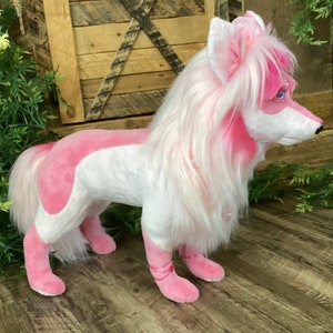 May include: A pink and white plush toy dog with long white fur and pink accents. The dog is standing on a wooden floor.