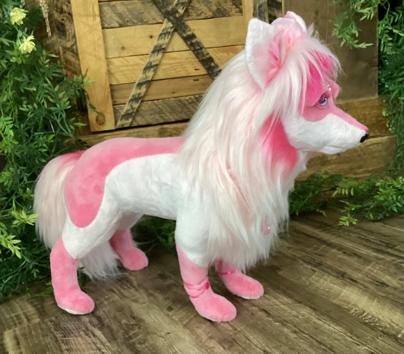 May include: A pink and white plush toy dog with long white fur and pink accents. The dog is standing on a wooden floor.