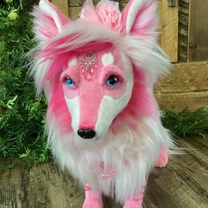 May include: A pink and white plush fox with a jeweled headpiece and a pink flower on its head. The fox is sitting on a wooden surface.