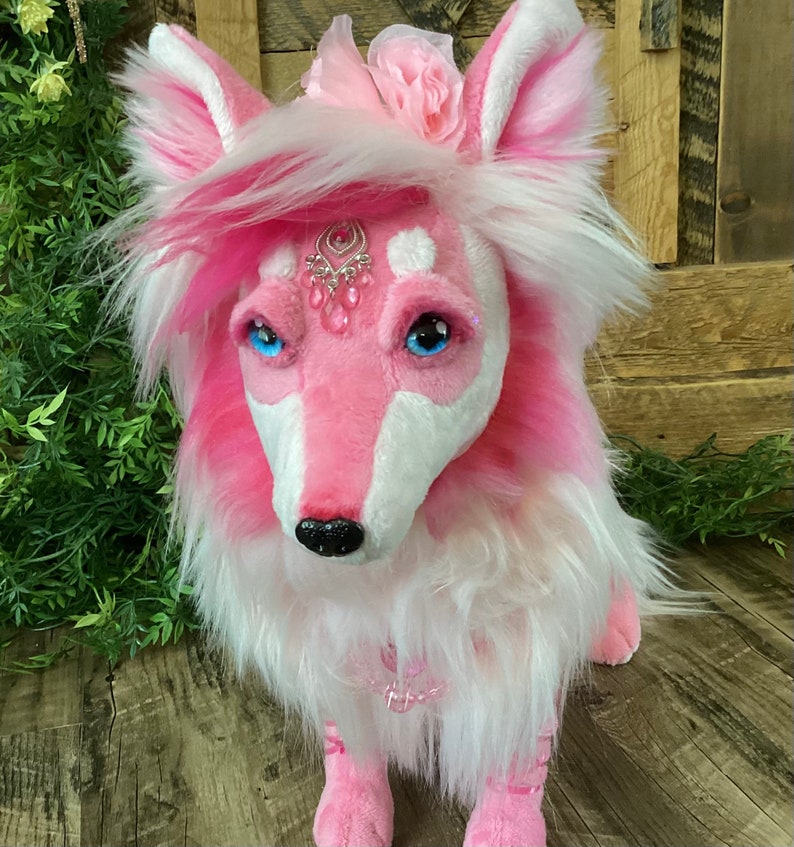 May include: A pink and white plush fox with a jeweled headpiece and a pink flower on its head. The fox is sitting on a wooden surface.